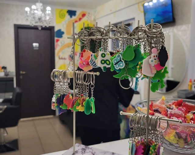 Suport cu brelocuri colorate pentru copii; în fundal se vede frizeria
